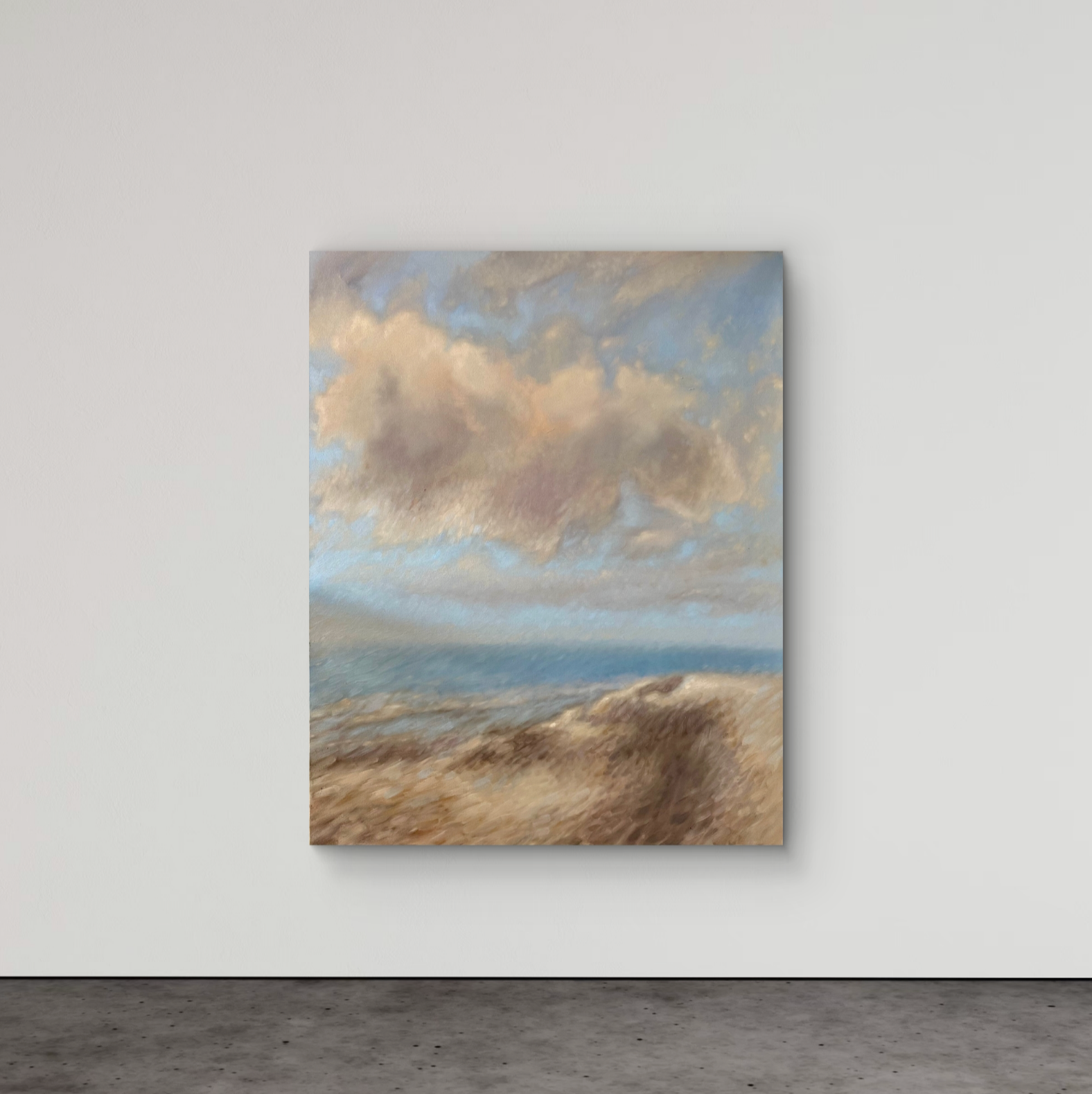 A vertical canvas painting of a beach landscape with soft blue sky, beige clouds, and sandy dunes, displayed on a light gray wall above a dark gray floor.
