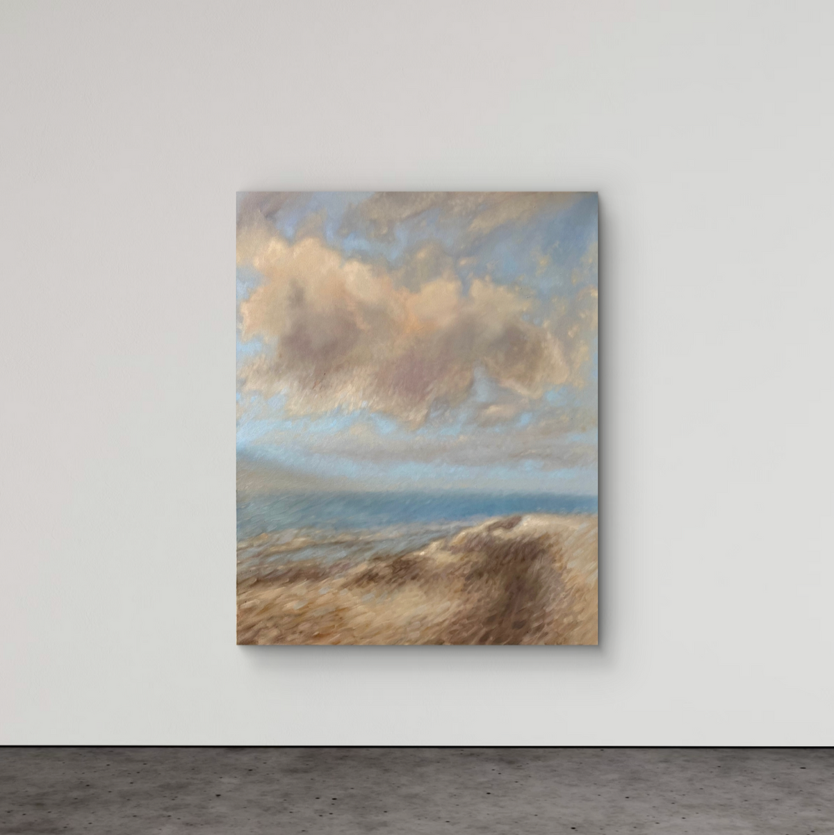 A vertical canvas painting of a beach landscape with soft blue sky, beige clouds, and sandy dunes, displayed on a light gray wall above a dark gray floor.